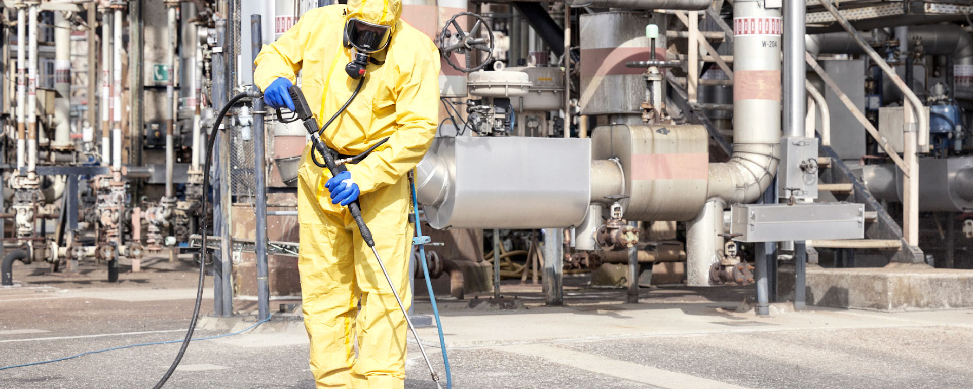 Ein REYM-Mitarbeiter in PSA mit gelbem Schutzanzug und Atemschutz führt Hochdruckreinigungsarbeiten vor einer Industrieanlage durch